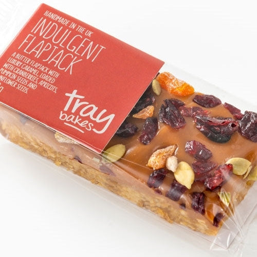 Packaged Traybakes Wrapped Indulgent Flapjack with visible caramel chunks, cranberries, apricots, and seeds in a clear plastic wrap.