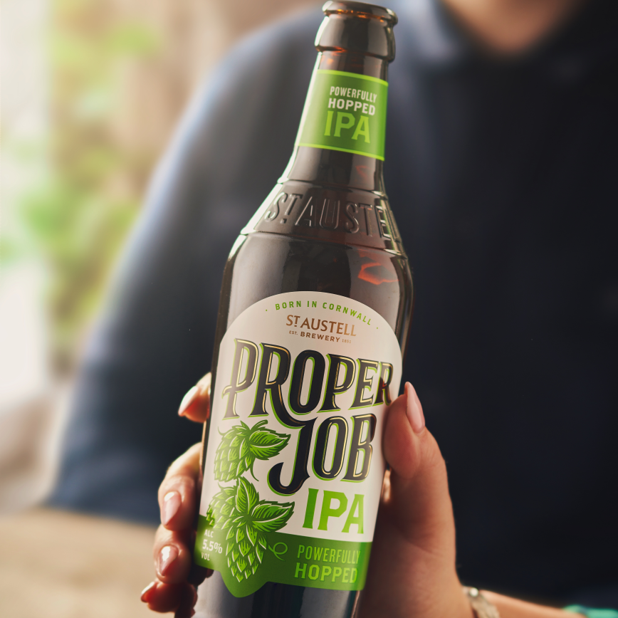 A hand holding a brown glass bottle of Proper Job IPA with a green and white label, in front of a blurred background.