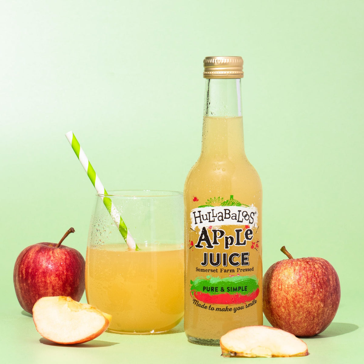 A glass bottle of Hullabaloos Apple Juice with a straw, accompanied by apples and apple slices on a green background.