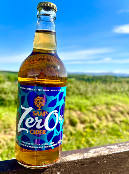 A bottle of Sams Zero Cider sits on a wooden railing with a blurred green landscape and blue sky in the background. The bottle is filled with amber-colored cider and features a blue and purple label with white and yellow text.