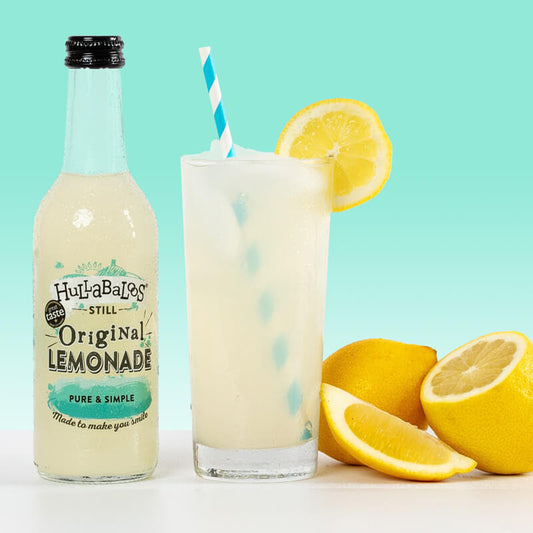 A bottle of HULLABALOOS Original Lemonade alongside a glass with lemonade and ice, garnished with a lemon slice, and accompanied by fresh lemons cut in half.