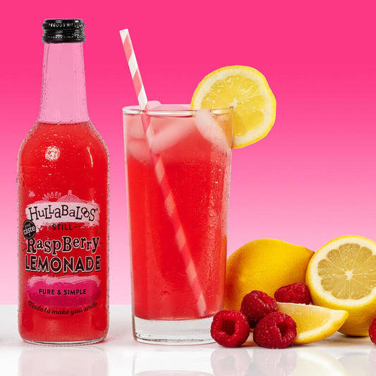 A glass of Hullabaloo's Still Raspberry Lemonade with a straw, accompanied by a bottle of the beverage and sliced raspberries and lemon wedges on a pink background.